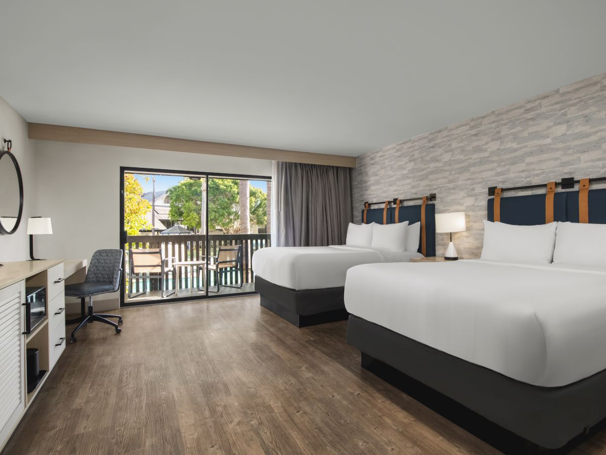 Modern hotel room with two beds, a large window/door, curtains, a desk, chair, and fireplace&mdash;sleek, bright, and tidy.