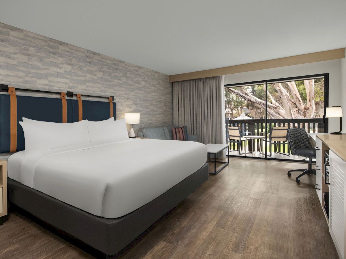 A modern hotel room with a large bed, lamps, a seating area, sliding doors, and a view of trees.