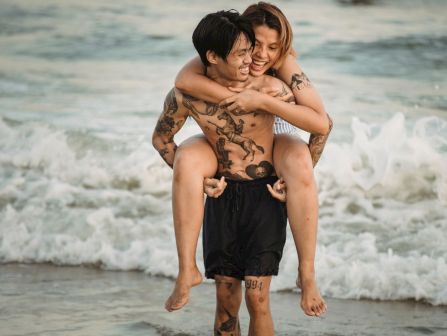 A tattooed couple hugs and laughs on a beach, standing in front of the waves in a warm, playful moment together.