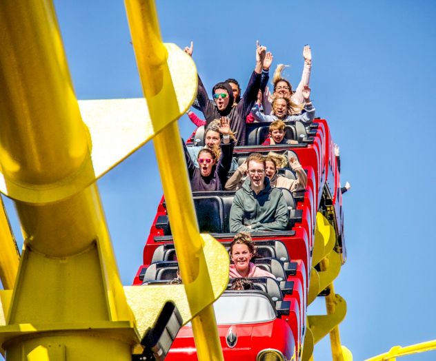 People ride a red roller coaster car, laughing and waving as they pass yellow supports in a sunny sky.