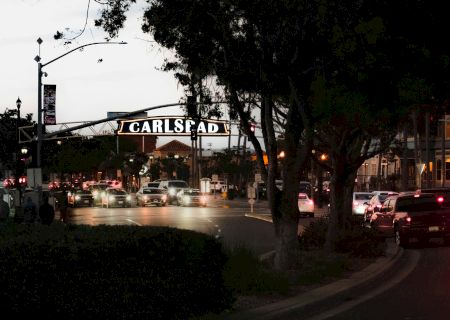 A busy evening street with cars and a glowing arch sign reading &ldquo;Carlsbad,&rdquo; framed by trees and traffic lights.