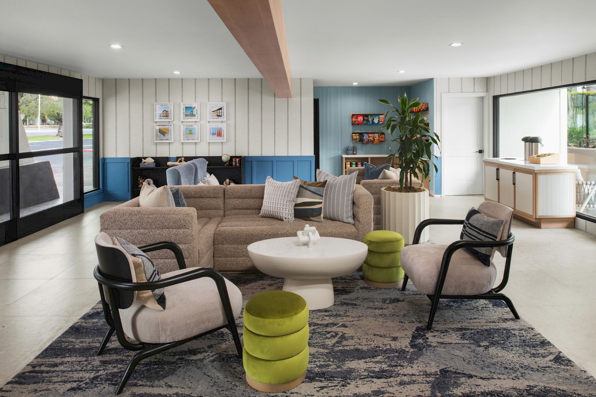 Bright modern lounge with beige sofa, round coffee table, green stools, shelves, framed photos, big windows, and indoor plant.