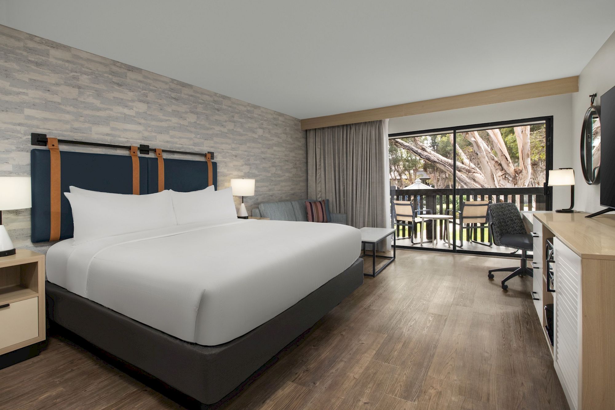 A modern hotel room with a large bed, lamps, desk, chairs, and floor-to-ceiling window showing an outdoor patio.