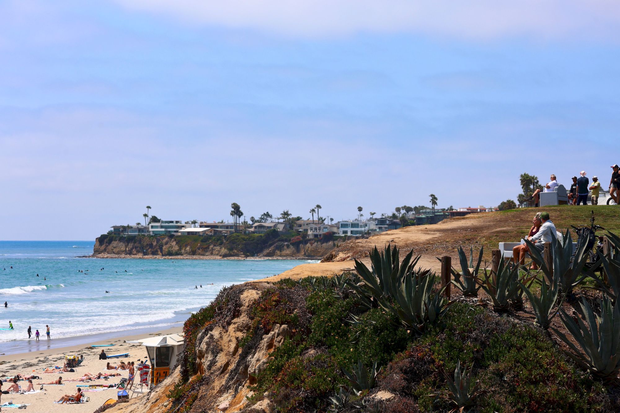 A sunny coastal viewpoint with beachgoers below, ocean waves, cliffs lined with agave plants, and people overlooking from the hilltop.