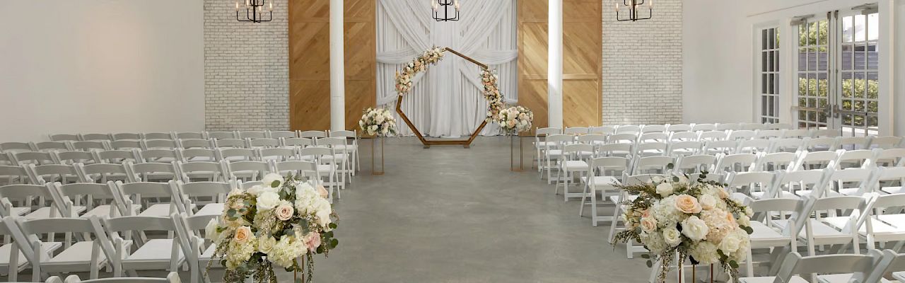 A bright wedding ceremony setup with rows of white chairs, floral arrangements, and hanging chandeliers over the aisle and altar.