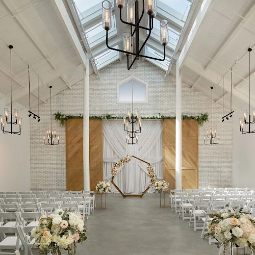 A bright wedding ceremony setup with rows of white chairs, floral arrangements, and hanging chandeliers over the aisle and altar.
