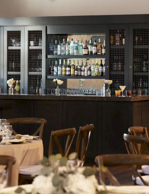 A restaurant bar with glass shelves of liquor, drink glasses, and dining table settings in the foreground, lit for evening service.