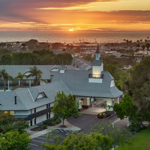 A coastal resort at sunset, with palm trees and grey-roof buildings overlooking the ocean horizon.