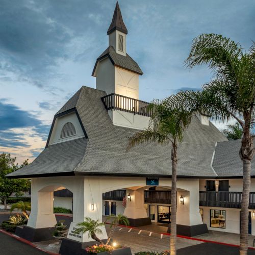 A white chapel with steeple and arched openings, lit at dusk, surrounded by palm trees and a parking lane.