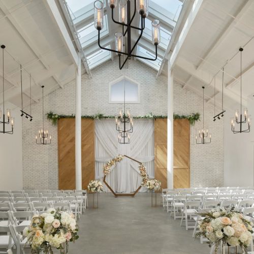 Bright church-style wedding hall with white chairs, floral aisle decor, a floral arch at the altar, and hanging chandeliers.
