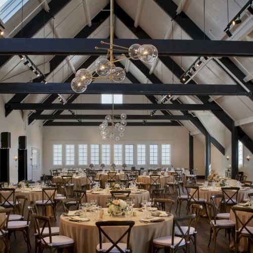A banquet hall set with round tables, gold chairs, and large crystal chandeliers hanging from a high ceiling.