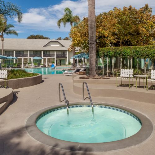 A sunny resort courtyard with a round hot tub, a swimming pool, lounge chairs, umbrellas, palm trees, and modern apartment buildings.