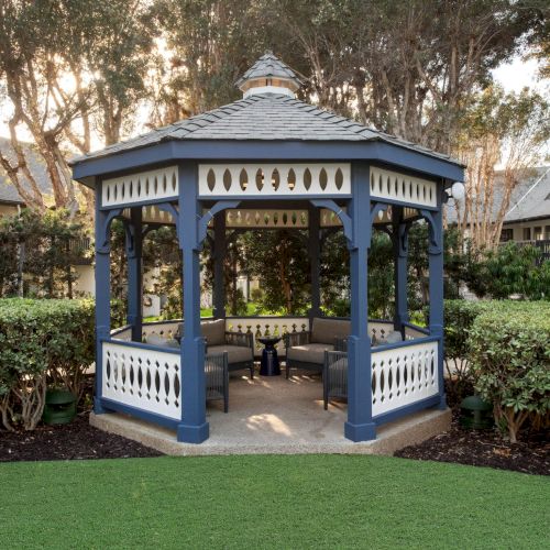 A blue outdoor gazebo with benches sits in a landscaped garden courtyard as tall trees glow in the golden sunset light.