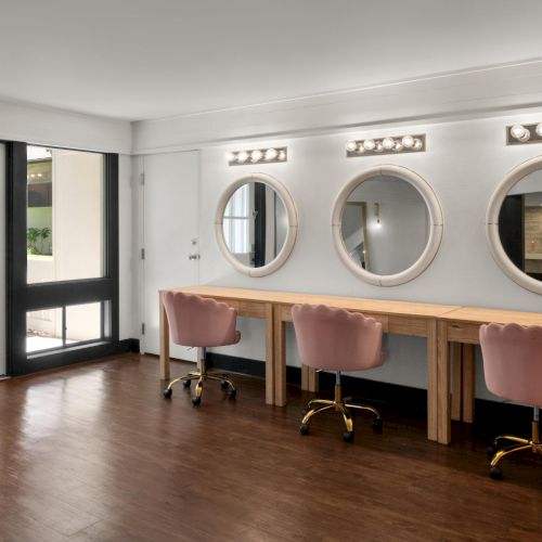 Modern office interior with a wooden desk, pink swivel chairs, round mirrors, and warm LED wall lights by large glass doors.