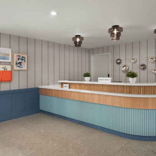 A modern hotel reception lobby with a curved blue-and-wood front desk, pendant lights, striped wallpaper, and framed art on the wall.