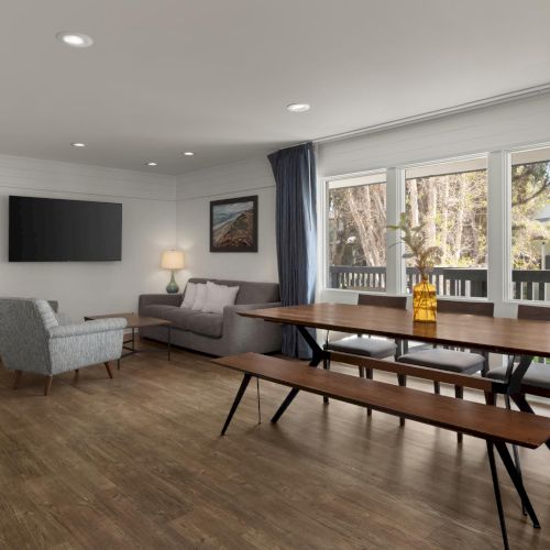 Bright living room with gray sectional, wooden dining table, benches, dark curtains, and large windows overlooking trees.