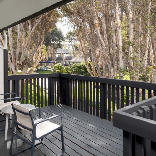 A quiet apartment balcony with patio table and chairs, overlooking a courtyard with tall trees and a distant umbrella.