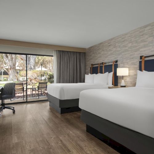 Bright hotel room with two white beds, lamps, and a large window showing trees outside.