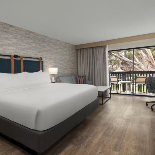 Modern hotel room: large bed, lamps, desk, and chairs, with curtains and a view outside through sliding glass doors.