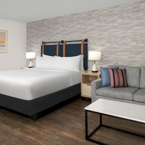 A modern hotel room with a king bed, lamps, framed wall art, and a gray couch by a stone-pattern accent wall.