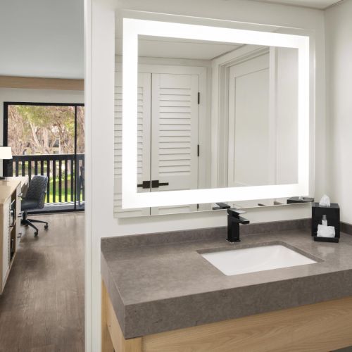 Modern hotel bathroom with double sink, dark faucet, and a TV in the adjacent room window.