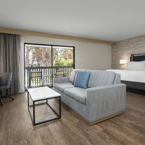 A modern hotel room with a king bed, gray sofa, desk, and large patio doors opening to a balcony.