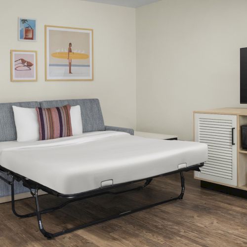 A hotel-style room with a bed and pillows, framed surf art, a flat-screen TV on a console, and a microwave cabinet beside it.