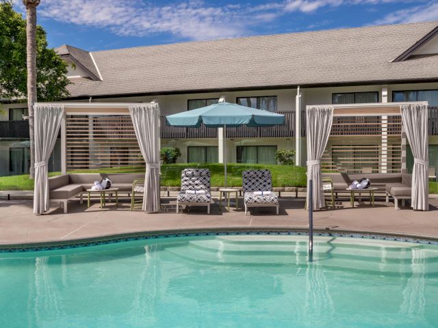 Sunny backyard patio with a turquoise pool, lounge chairs, and a covered cabana with curtains in front of modern homes.