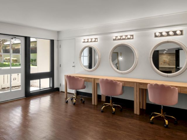 Bright office interior with a wooden desk, pink swivel chairs, and three round lit mirrors on a white wall, plus patio doors.
