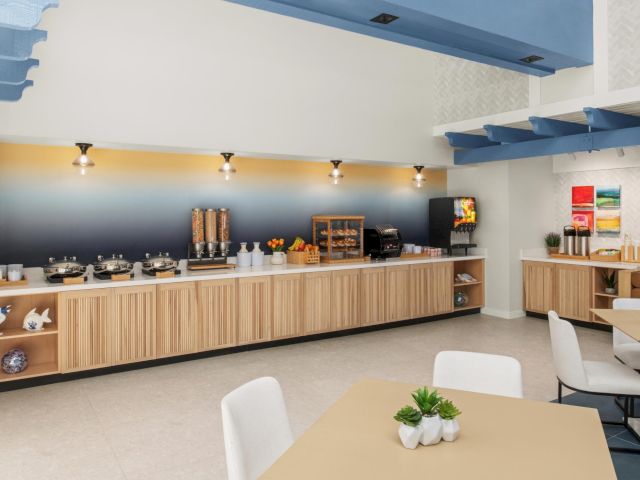 Modern buffet station with coffee dispensers, fruit, a grill, and colorful wall art in a bright lounge.