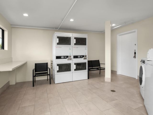 A bright laundry room with stacked white washer-dryers, a folding bench, and a sink/cabinet on the right.